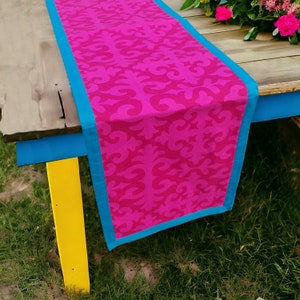 May include: A pink table runner with a blue border and a swirling pattern. The runner is on a wooden table with a blue and yellow painted leg.