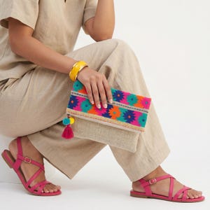 May include: A beige linen clutch bag featuring a fold-over flap with a vibrant geometric pattern in orange, pink, teal, and dark blue. The bag includes a decorative tassel with two small pom-poms attached to the side zipper pull.