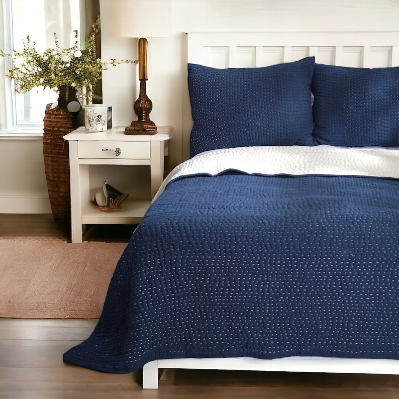 May include: A navy blue quilted bedspread and two matching pillow shams. The bedspread features a white underside and is placed on a white bed frame. The room includes a white nightstand, a lamp, and a decorative vase.