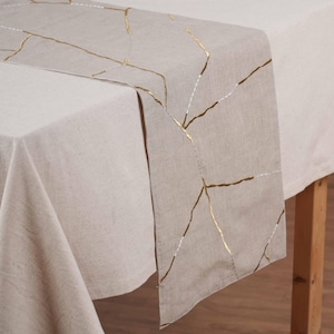 May include: Beige linen table runner with gold and white stitched geometric design. The runner is draped over a beige tablecloth, creating a layered effect. The table is made of wood.
