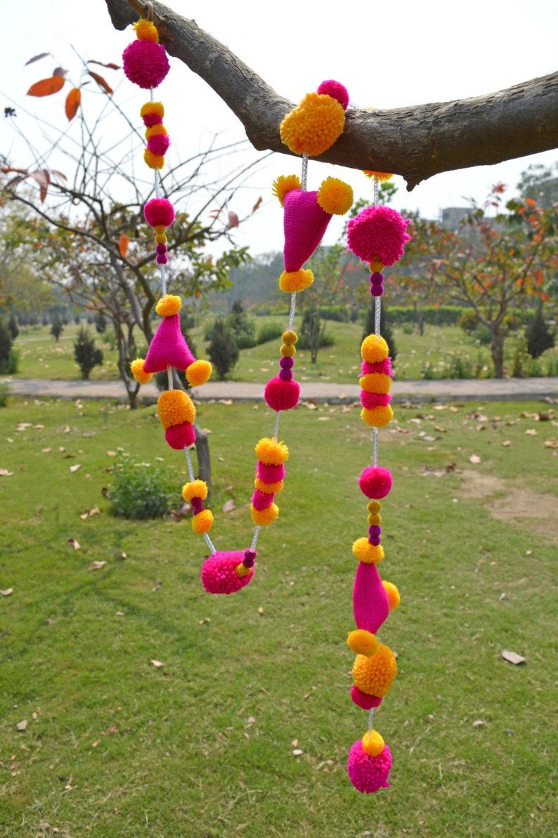 May include: A garland of pink and yellow pom-poms hangs from a tree branch. The garland is made of yarn and is strung together with white thread. The pom-poms are arranged in a repeating pattern of pink, yellow, and pink.