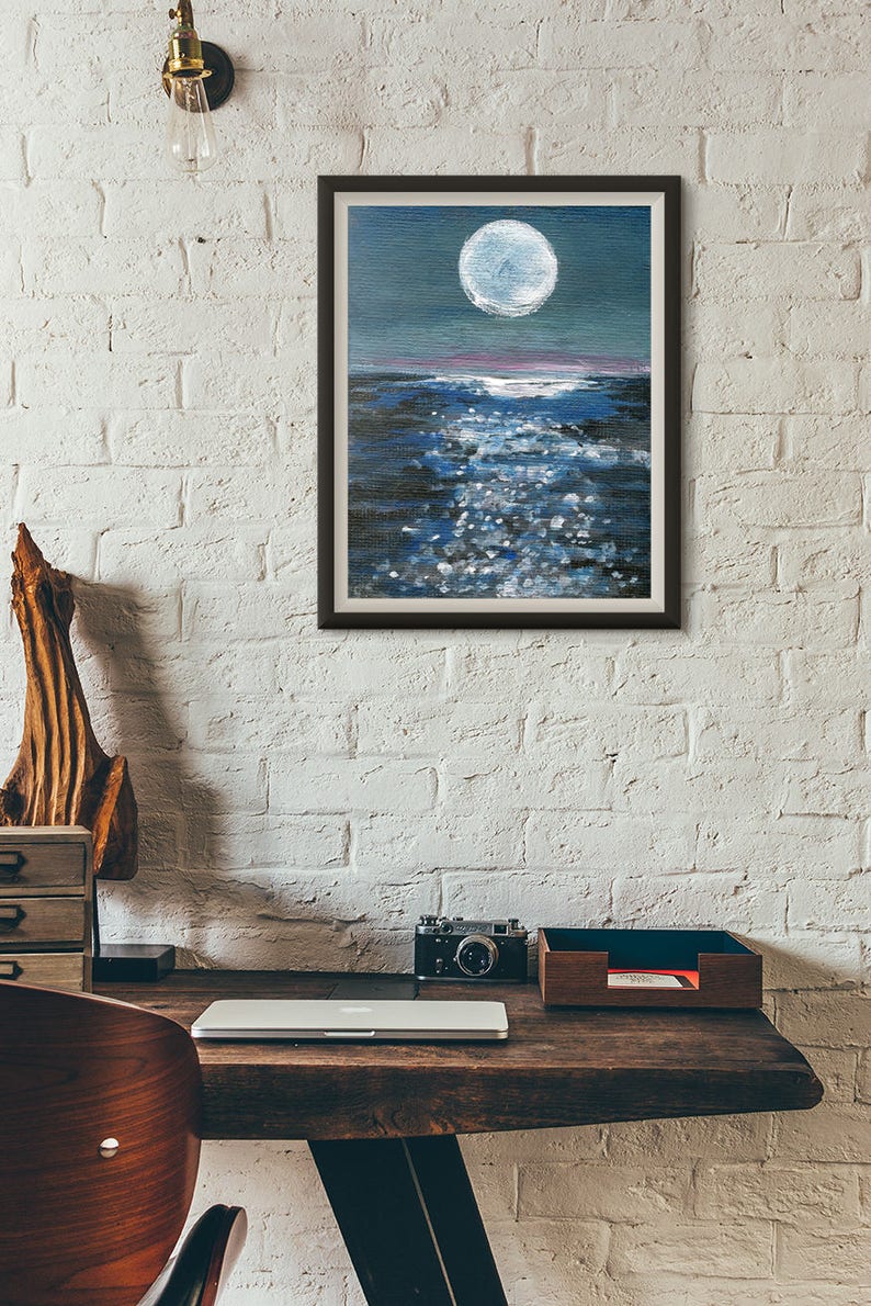 May include: Framed painting of a moonlit ocean scene. The artwork features a large, bright white moon in a dark blue sky, reflecting on the water. The water is a mix of dark and light blues with white highlights, creating a shimmering effect.