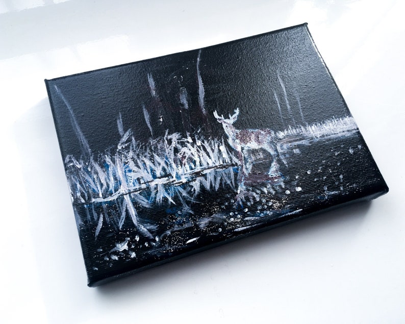 May include: A small, rectangular canvas painting depicting a deer in a dark forest scene. The artwork features a deer with white spots, standing near a body of water. The painting uses white, blue, and black paint to create a nighttime landscape.