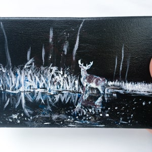 May include: An original painting on canvas depicting a deer standing in water at night. The artwork features a dark background with white and blue accents, creating a reflective water effect. The deer is painted with white and brown tones.
