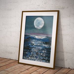 May include: A framed painting depicts a nighttime seascape. A large, bright white moon dominates the upper portion of the canvas, above a dark blue ocean with white highlights. The frame is a warm brown color, and the painting is leaning against a white wall.