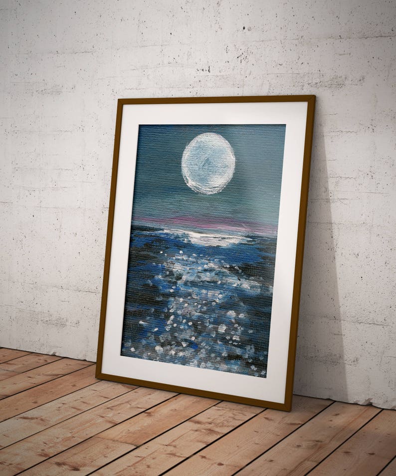 May include: A framed painting depicts a nighttime seascape. A large, bright white moon dominates the upper portion of the canvas, above a dark blue ocean with white highlights. The frame is a warm brown color, and the painting is leaning against a white wall.