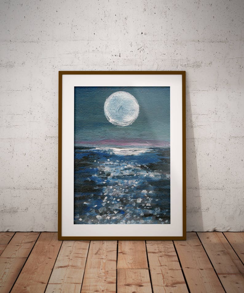 May include: Framed painting of a moonlit ocean scene. The artwork depicts a large, white moon in a dark blue sky above dark blue water with white highlights. The painting is in a brown frame and leans against a white wall.