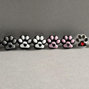 May include: A collection of paw print stud earrings in various colors, including gray, white, and pink. Each paw print features a heart in the center. The earrings are displayed on a light gray surface.