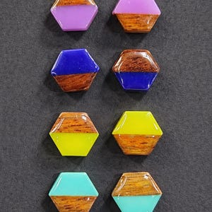 May include: Eight hexagonal wooden and resin earrings in various colors. The top half of each earring is a different color of resin: purple, blue, yellow, and teal. The bottom half is wood grain. The earrings are arranged on a dark gray surface.