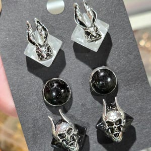 May include: A set of four pairs of earrings on a black card. The earrings feature silver-toned rabbit skull studs, black round studs, and silver-toned devil skull studs. The rabbit skulls are on white square bases.