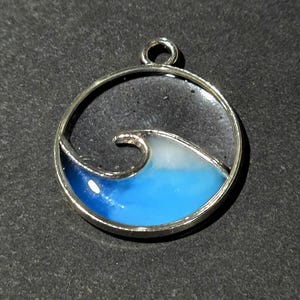 May include: Silver-toned pendant featuring a stylized wave design. The wave is depicted in shades of blue and white, encased within a circular frame. The pendant has a loop at the top for a chain.