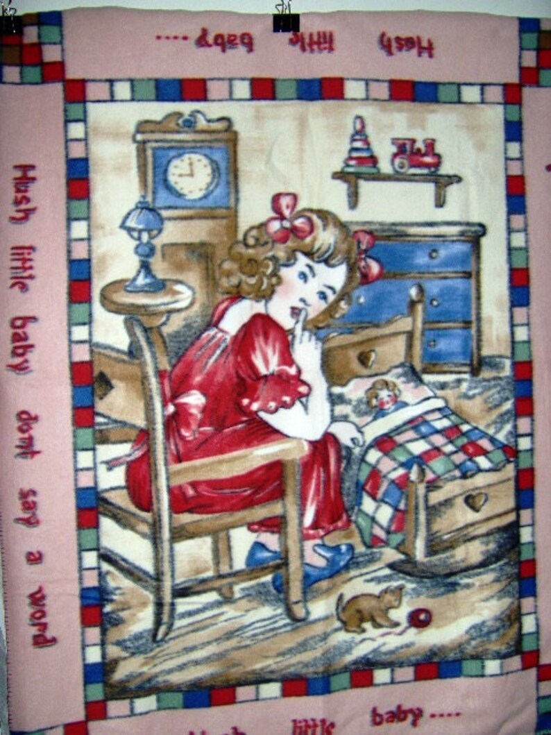 May include: A pink and cream colored blanket with a vintage illustration of a girl in a red dress sitting in a rocking chair. The girl is holding her finger to her lips and looking at a doll in a crib. The text "Hush little baby don't say a word" is printed on the left side of the blanket.
