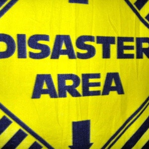 May include: A yellow diamond-shaped sign with black stripes and an arrow pointing down. The sign reads "DISASTER AREA" in bold blue letters.