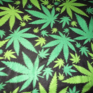 May include: A black fabric with a repeating pattern of green and light green cannabis leaves. The leaves vary in size and are scattered across the surface, creating a bold, eye-catching design. The fabric appears to be soft and possibly fleece.