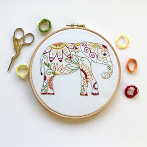 May include: Embroidered hoop art featuring a white elephant with colourful floral patterns in shades of red, yellow, green, and orange. The elephant is surrounded by a wooden hoop.