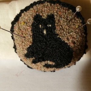 May include: A round pincushion with a black cat design. The pincushion is made of brown fabric and has a textured surface. There are three pins with white pearl heads sticking into the pincushion.