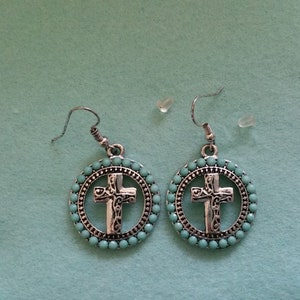 May include: A pair of silver dangle earrings with a turquoise cross design. The earrings have a silver hook and a round turquoise bead design.