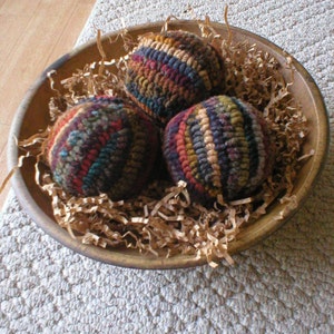 May include: Three colorful yarn balls in a brown bowl with brown shredded paper. The yarn balls are made of different colors of yarn, including red, blue, green, yellow, and brown.