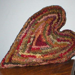 May include: A heart-shaped rug with a patchwork design. The rug features a variety of colors, including red, green, and brown. The rug is made of fabric strips sewn together in a spiral pattern. The rug is placed on a wooden surface.