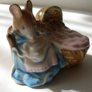 May include: A ceramic figurine of a brown mouse holding a wicker basket with a pink blanket inside. The mouse is wearing a blue and white dress with pink polka dots.