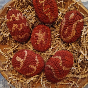 May include: Six red fabric eggs with white stitching in a decorative nest of brown paper shreds. The eggs have a textured surface and are arranged in a circular pattern.
