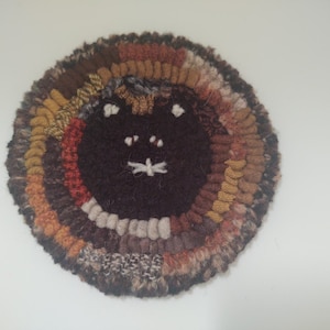 May include: A round, brown, orange, and beige wool rug with a brown cat face in the center. The cat face has white eyes and a white nose.