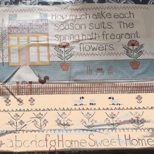 May include: A beige cross-stitch sampler with a house, flowers, and a fence. The text reads "How much alike each season suits. The Spring hath fragrant flowers." and "Home Sweet Home".