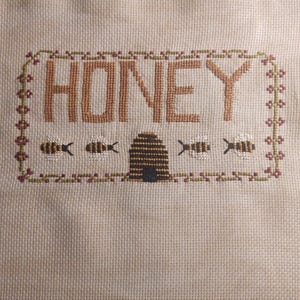 May include: Cross-stitch embroidery with the word "HONEY" in brown thread, a beehive in black and brown thread, and four bees in yellow and black thread. The embroidery is framed with a green and brown border.