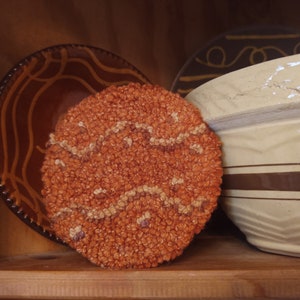 May include: A close-up of a brown and cream-colored woven rug with a wavy pattern. The rug is sitting on a wooden shelf with a brown and white ceramic bowl behind it.