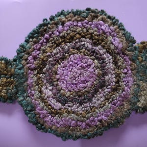 May include: A handmade, round rug with a floral design. The rug features concentric circles of varying colors, including purple, green, and brown. The rug has a textured, looped appearance and is set against a solid purple background.