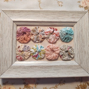May include: A framed wall art piece with eight colorful fabric rosettes in a white frame. The rosettes are arranged in two rows of four. The rosettes are made of different fabrics and have different patterns.