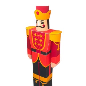 May include: A red and gold papercraft nutcracker soldier with a black mustache and a gold crown. The nutcracker is standing with its arms at its sides.