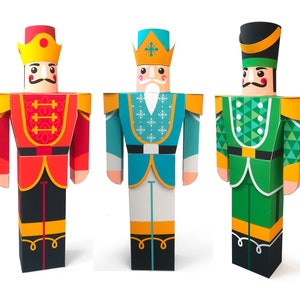 Puede incluir: Tres figuras de cascanueces de papel. El cascanueces de la izquierda es rojo con una corona amarilla y botas negras. El cascanueces del medio es azul con una corona blanca y botas blancas. El cascanueces de la derecha es verde con un sombrero negro y botas negras.