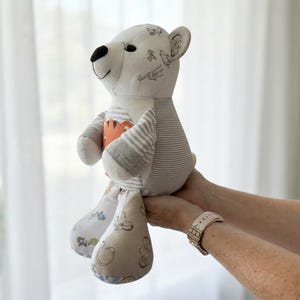 May include: A handmade plush bear with a white body, black eyes and nose, and patterned accents. The bear has striped arms and legs, and an orange and white chest. The bear is being held up by a person.