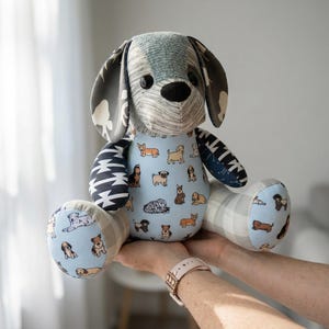 May include: A handmade plush dog toy with a patchwork design. The dog's head and ears are patterned with gray and blue fabrics, while the body features a light blue fabric with various dog illustrations. The paws and legs have a light blue background with dog illustrations.
