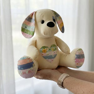 May include: A cream-coloured stuffed dog with black button eyes and a black nose. The dog's ears, paws, and a heart-shaped patch on its chest feature colourful patchwork fabric. A yellow button is sewn onto the heart.