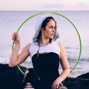 May include: A woman wearing a white and black hooded top and black ripped leggings, holding a green hula hoop. She is sitting on a dark rock formation near the ocean, with a cloudy sky in the background.