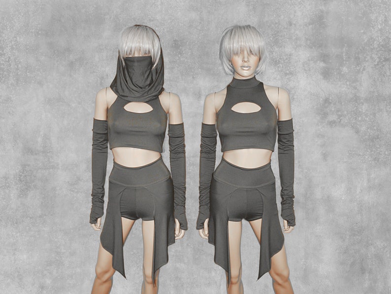 May include: Two mannequins wearing a black crop top with a keyhole cutout, black shorts with a long, flowing skirt overlay, and black fingerless gloves. The mannequins have short, silver hair.