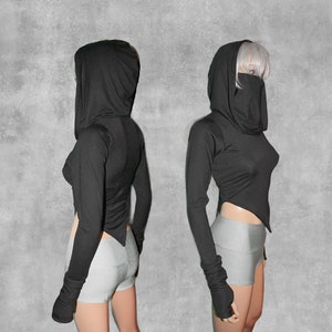 May include: Black long-sleeved hooded top with a high neckline and a long, flowing hood. The top is cropped and has a slightly asymmetrical hemline. The sleeves are long and have thumbholes. The top is made of a soft, stretchy fabric.