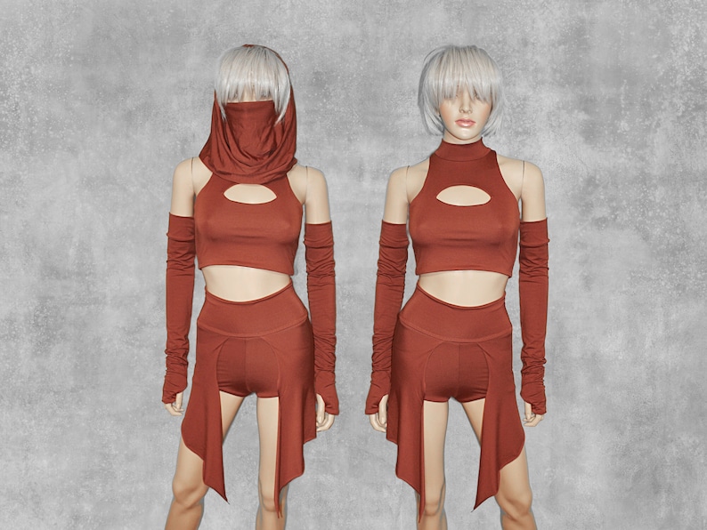 May include: A rust-colored outfit with a crop top, shorts, and a long skirt. The outfit has a keyhole cutout in the front of the crop top and a high neck. The outfit is worn by a mannequin with a white wig.