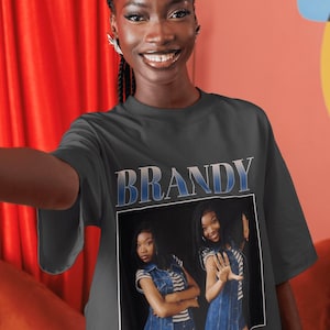 Pode incluir: Camiseta cinza escuro com o nome "BRANDY" em letras azuis e brancas. A camiseta exibe uma foto de uma mulher usando um colete jeans sobre uma camisa listrada. A mulher está sorrindo e posando.