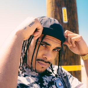 May include: A man with braided hair is wearing a black beanie and a tie-dye shirt with a circular patch. He has a tattoo on his wrist that says "God".