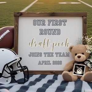 May include: An announcement with a football theme. The sign says "OUR FIRST ROUND DRAFT PICK JOINS THE TEAM APRIL 2026." There's a football, a white helmet, a teddy bear with an ultrasound image, and flowers.