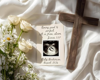 May include: A wooden cross rests on a white fabric, next to a card with an ultrasound image and text that reads "Every good & perfect gift is from above James 1:17" and "Baby Anderson August 2026". White roses and daisies are arranged nearby.