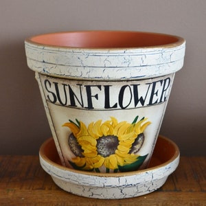 May include: A white ceramic planter with a distressed finish, featuring a painted sunflower design and the word "SUNFLOWER" in black lettering. The planter has a matching saucer.