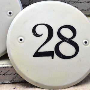 May include: White ceramic house number plaque with black painted number 28. The plaque has two small holes on the side.