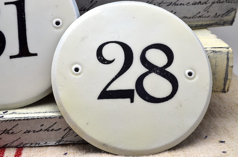 May include: White ceramic house number plaque with black painted number 28. The plaque has two small holes on the side.