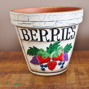 Puede incluir: Maceta de cerámica blanca con un acabado desgastado. La maceta tiene una inscripción negra que dice "BERRIES" y un diseño pintado de fresas, arándanos y frambuesas.
