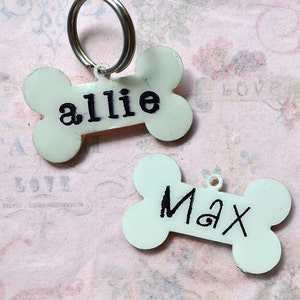 May include: Two white bone-shaped dog tags with black lettering. The top tag says "allie" and the bottom tag says "Max".