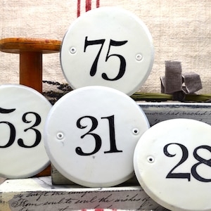 May include: Five white ceramic round and oval signs with black numbers. The numbers are 75, 53, 31, and 28.